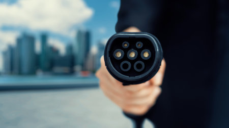 Focus EV charger pointing in front of camera with businessmans hand in blurry background of cityscape and cloud. Electric car charger plug and alternative clean energy reducing CO2 emission.Peruseの写真素材