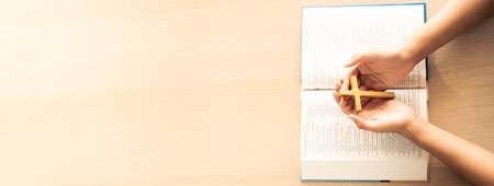 Female god believer holding wooden cross on opened holy bible book at light wooden church table. Top view. Concept of hope, religion, faith, christianity and god blessing. Warm background. Burgeoning.の写真素材