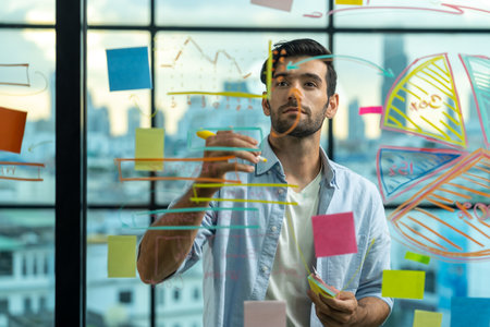 Portrait of young handsome businessman writing marketing idea by using colorful marker on glass wall...