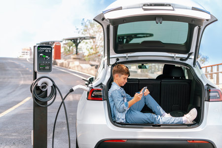 Little boy sitting on car trunk, using smartphone while recharging eco-friendly car from EV charging station. EV car road trip travel as alternative vehicle using sustainable energy concept. Perpetualの写真素材