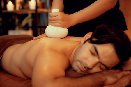 Hot herbal ball spa massage body treatment, masseur gently compresses herb bag on man body. Tranquil and serenity of aromatherapy recreation in warm lighting of candles at spa salon. Quiescentの写真素材