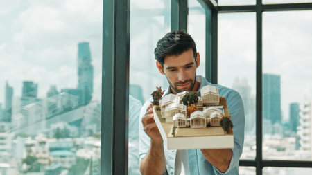 Businessman in casual outfit holding house model while checking house construction. Architect engineer inspect building model while standing near window with skyscraper. Engineering. Traceryの写真素材