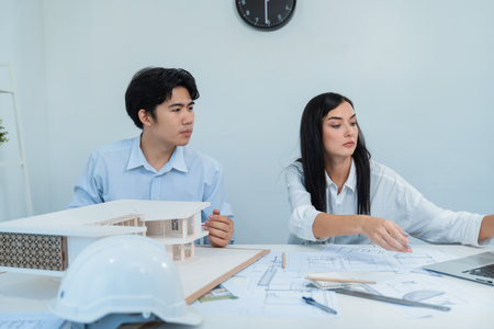 Professional male asian architect using ruler to measure house model length while young beautiful caucasian colleague using laptop to analyzed data on meeting table with house model. Immaculate.の写真素材