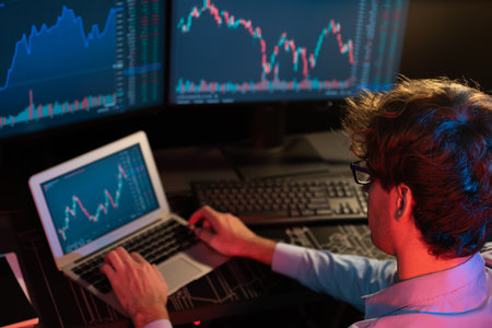 Young investor stock trader searching on laptop comparing with dynamic exchange rate screens of...