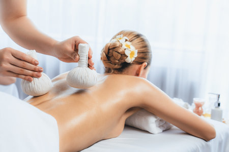 Hot herbal ball spa massage body treatment, masseur gently compresses herb bag on woman body. Tranquil and serenity of aromatherapy recreation in day lighting ambient at spa salon. Quiescentの写真素材