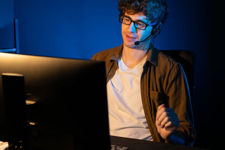 Young creative businessman communicating with listeners on laptop screen or meeting with team member, wearing headphones at neon light modern home office. Concept of working at night time. Gusher.の写真素材
