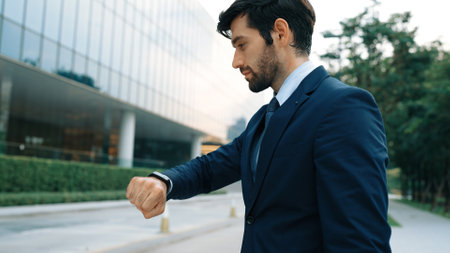 Skilled caucasian business man looking at time while standing at building. Attractive executive manager using smart watch while waiting for partner. Time management, punctuality concept. Exultant.の写真素材