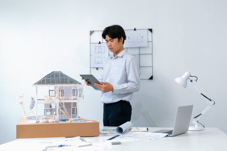 Professional handsome asian project manager using tablet to check house model while inspect house model structure carefully at office with blueprint hanged behind. Creative design. Immaculate.の写真素材