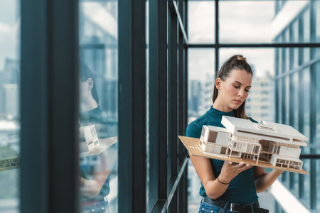 Beautiful young architect engineer holds architectural model while inspect house model. Professional interior designer checking house construction while standing near window with city view. Tracery.の写真素材