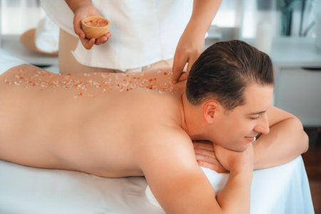 Blissful man customer having exfoliation treatment in luxury spa salon with warmth candle light ambient. Salt scrub beauty treatment in health spa body scrub. Quiescentの写真素材