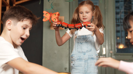 Girl playing wooden train while diverse student sitting at play room. Boy looking at lovely cute student hold toy while his friend talking about lesson in break time. Creative activity. Erudition.の写真素材