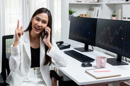 Young beautiful Asian businesswoman talking to broker on phone with looking on pc screen to analyze...