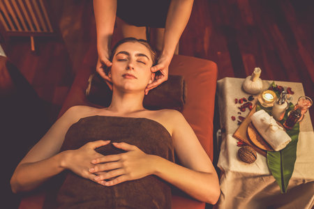 Caucasian woman enjoying relaxing anti-stress head massage and pampering facial beauty skin recreation leisure in warm candle lighting ambient salon spa in luxury resort or hotel. Quiescentの写真素材