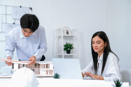 Professional male asian architect using ruler to measure house model length while young beautiful caucasian colleague using laptop to analyzed data on meeting table with house model. Immaculate.の写真素材