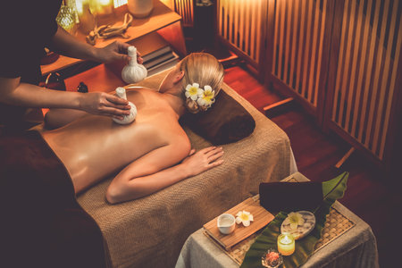 Hot herbal ball spa massage body treatment, masseur gently compresses herb bag on woman body. Tranquil and serenity of aromatherapy recreation in warm lighting of candles at spa salon. Quiescentの写真素材