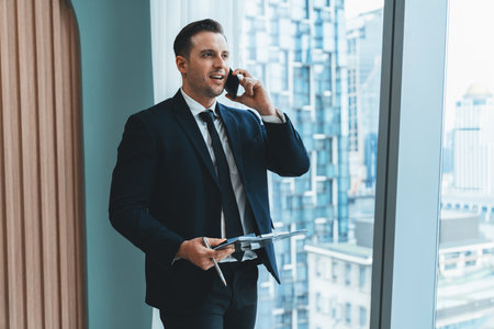Portrait of professional businessman stand near window with skyscraper while phone calling and holding laptop. Busy executive manager holding laptop while multitasking working. Closeup. Ornamented.の写真素材