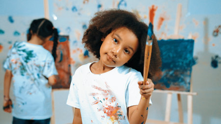 Cute african girl holding painted brush while student drawing canvas behind. Multicultural children...
