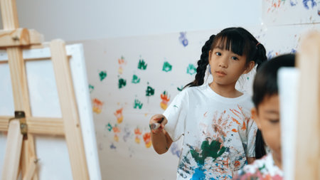 Playful student painted or draw canvas at stained wall in art lesson. Asian girl wearing white shirt with stained color while standing at stained wall with hand print. Creative activity. Erudition.の写真素材