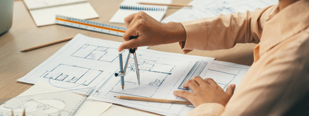 Professional architect drawing blueprint by using divider on the table with stationary and architectural document scatter around at architectural office. Closeup. Focus on hand. Delineation.の写真素材