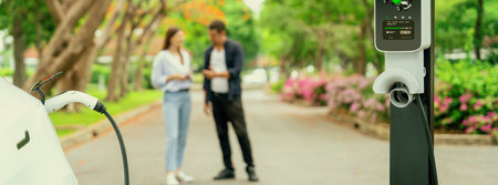 Focused EV car recharging battery on blurred background of lovey couple during autumnal road trip vacation by electric vehicle recharging battery. Eco friendly travel during autumn. Panorama Exaltの写真素材