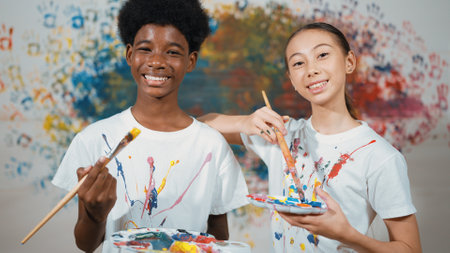 Highschool student with mixed races standing and looking at camera at art lesson. Diverse children hold the paintbrush while pose with confident at colorful stained wall. Creative style. Edification.の写真素材