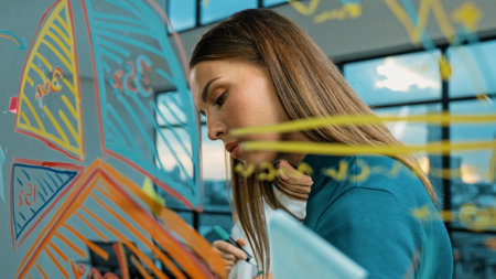 Smart businesswoman drawing graph while looking marketing plan at glass wall. Professional female...