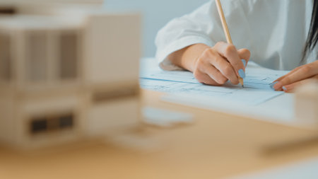 Closeup of beautiful young architect engineer hand drawing, drafting blueprint with blueprint and house model placed on working table at modern office. Focus on hand. Blurring background. Immaculate.の写真素材