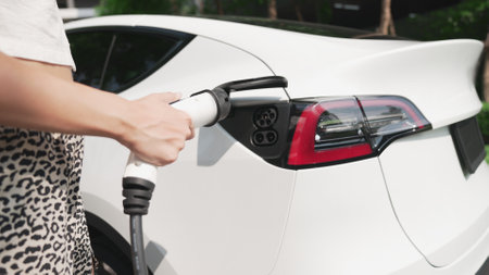 Young woman travel with EV electric car charging in green sustainable city outdoor garden in summer....