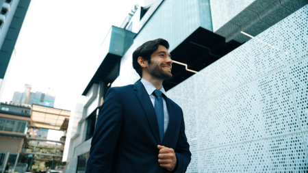 Smart business man wearing suit and smiling while walking at street near white modern architectural building. Attractive handsome manager looking skyscraper in urban city. Copy space. Exultant.の写真素材