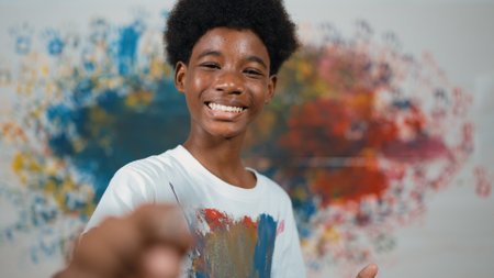 African boy standing at colorful stained wall while holding brush and point at camera. Happy highschool student wearing white shirt with stain color and look at camera with confident. Edification.の写真素材