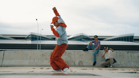Hip hop man wearing headphone while pose at camera with confident at bridge. Active energetic hipster perform freestyle dance or footstep while friend cheering behind. Outdoor music 2024. Sprightly.の写真素材