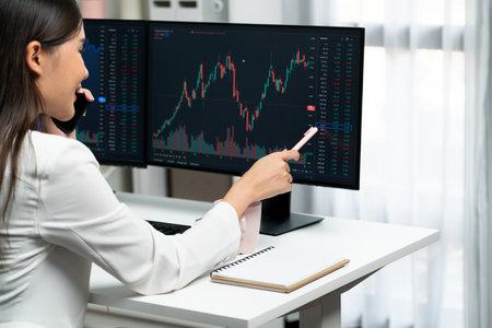 Young beautiful Asian businesswoman calling to broker, pointing with pen in increased exchange stock...