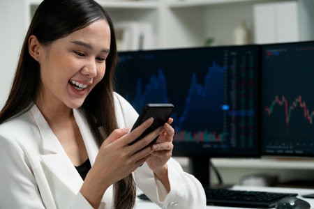 Smiling young Asian businesswoman with happy face looking on smartphone in profitable of exchange stock market graph at modern office. Concept of investing high profit in wealth security. Stratagem.の写真素材