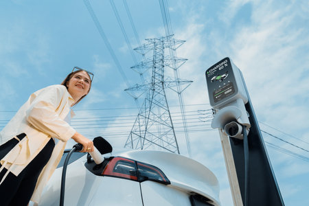 Woman recharge EV electric car battery at charging station connected to electrical power grid tower on sky background as electrical industry for eco friendly vehicle utilization. Expedientの写真素材