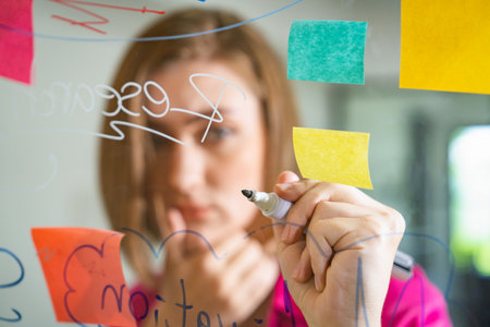 Closeup of young beautiful caucasian business leader presents marketing idea while writing marketing idea on glass board with mind map and colorful sticky notes. Portrait. Brainstorm. Immaculate.の写真素材