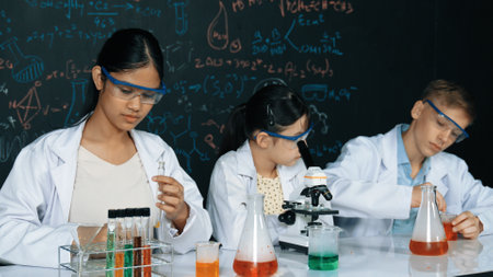 Cute girl looking under microscope while student doing experiment at blackboard with theory written. Young scientist inspect colored solution at table with experimental equipment placed. Edification.の写真素材