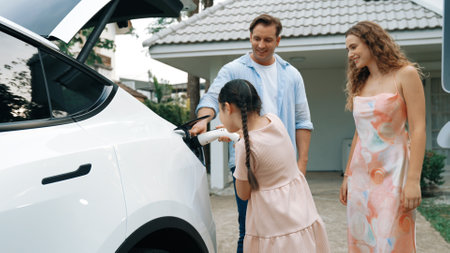 Happy little young girl learn about eco-friendly and energy sustainability as she help her family recharge electric vehicle from home EV charging station. EV car and modern family concept. Synchronosの写真素材