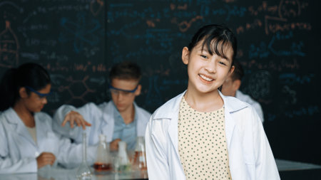 Cute asian scientist smile to camera while diverse group doing experiment. Young happy schoolgirl standing at blackboard with chemical theory written while wearing lab coat at laboratory. Edification.の写真素材