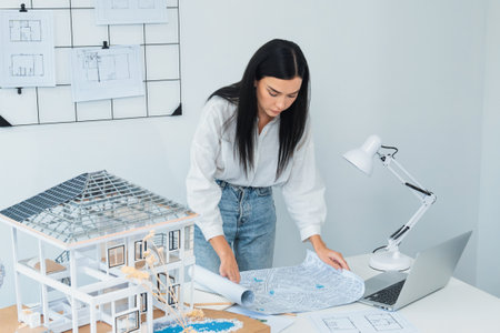 Portrait of Young beautiful caucasian architect focus on comparing house structure between house model and blueprint and laptop placed on meeting table. Check, inspect, estimate concept. Immaculate.の写真素材