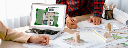 Professional architect represented ECO house displayed on laptop to project manager on messy table with wooden block, blueprint, architectural document placed at modern office. Close up. Delineation.の写真素材