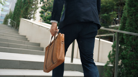 Back view of caucasian manager with formal suit walking up stairs while holding his suitcase in...