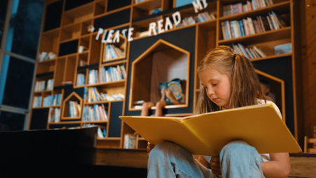 Young smart caucasian girl picking reading a book while sitting at library. Clever child learning, studying, open a books at library. Attractive kid turning page with blurring background. Erudition.の写真素材