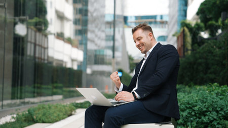 Caucasian project manager working on his laptop and celebrate for successful work while sitting at bench in green urban city. Businessman writing report and discussing idea with business team. Urbane.の写真素材
