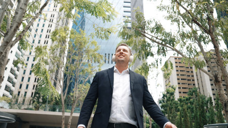 Business people wear headphone to listen relaxing music while walking workplace at green city. Low angle camera of manager move to song while go to meeting surround with tree and skyscraper. Urbane.の写真素材