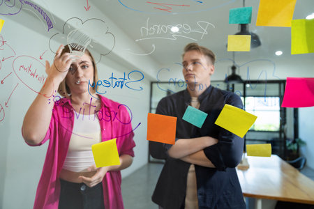 Couple of businesspeople discussing and brainstorming about solving marketing problem in front of glass board at business meeting by using mind map and sticky notes. Working together. Immaculate.の写真素材