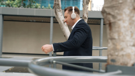 Happy project manager with headphone walking workplace while moving to relaxed music. side view of smart caucasian businessman going to meeting while dancing and walking along the city street. Urbane.の写真素材