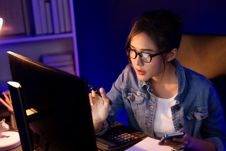 Thinking young beautiful Asian creative woman focusing on smartphone of sales target report business market graph comparing with report on computer at neon light modern office at night. Stratagem.の写真素材