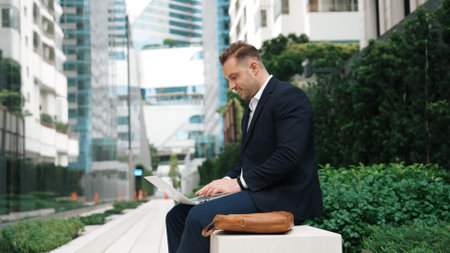 Manager working on his laptop and writing report to smart project manager in green urban city. Businessman looking analysis data and brainstorm idea by using computer while sitting at bench. Urbane.の写真素材