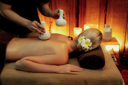 Hot herbal ball spa massage body treatment, masseur gently compresses herb bag on woman body. Tranquil and serenity of aromatherapy recreation in warm lighting of candles at spa salon. Quiescentの写真素材