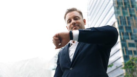 Low angle camera of businessman looking at watch to remind about time. Smart caucasian businessman checking time for being on time in important appointment or meeting with modern skyscraper. Urbane.の写真素材